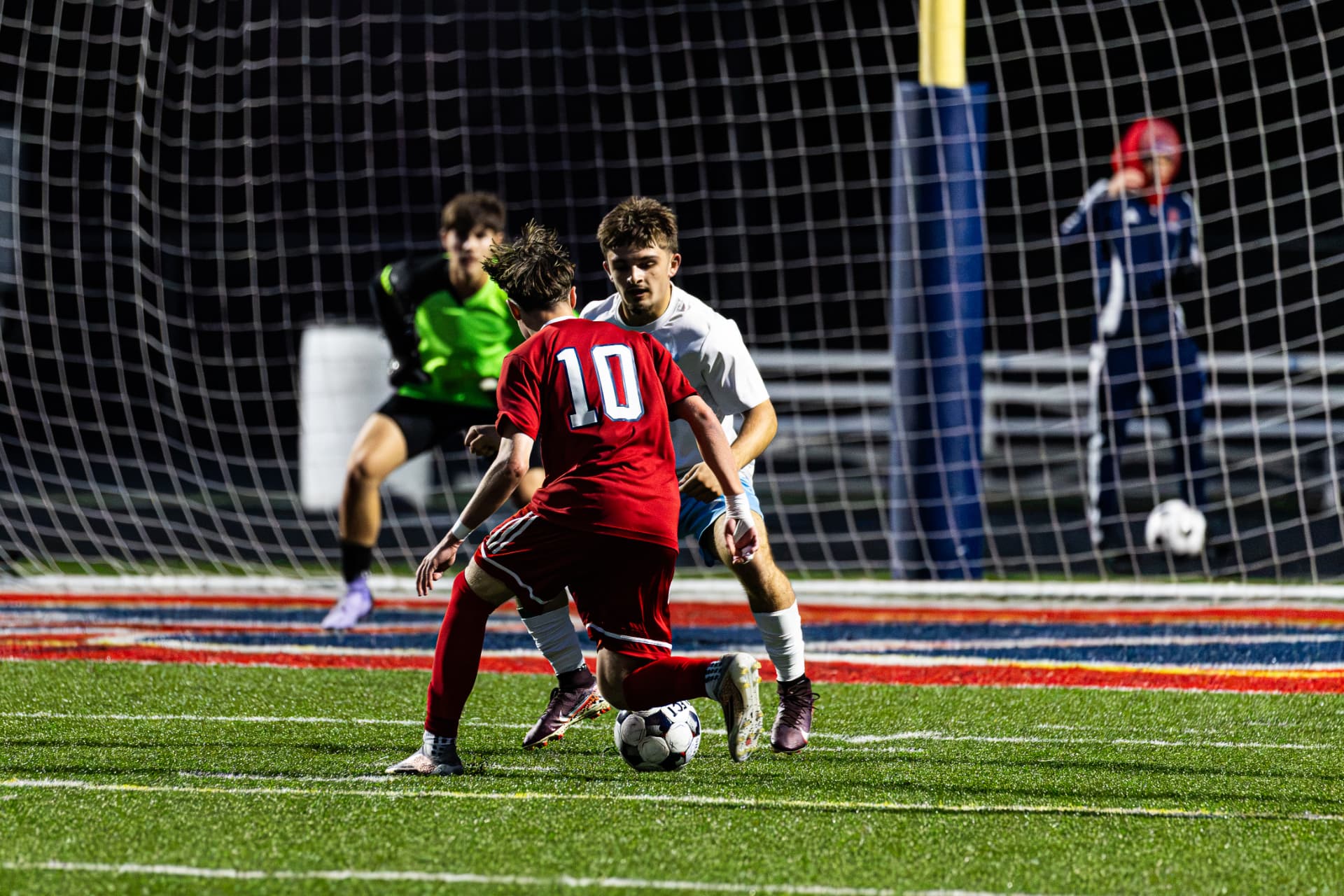 High school soccer players competing for the ball in front of the goal net at night — soccer photography by Striide